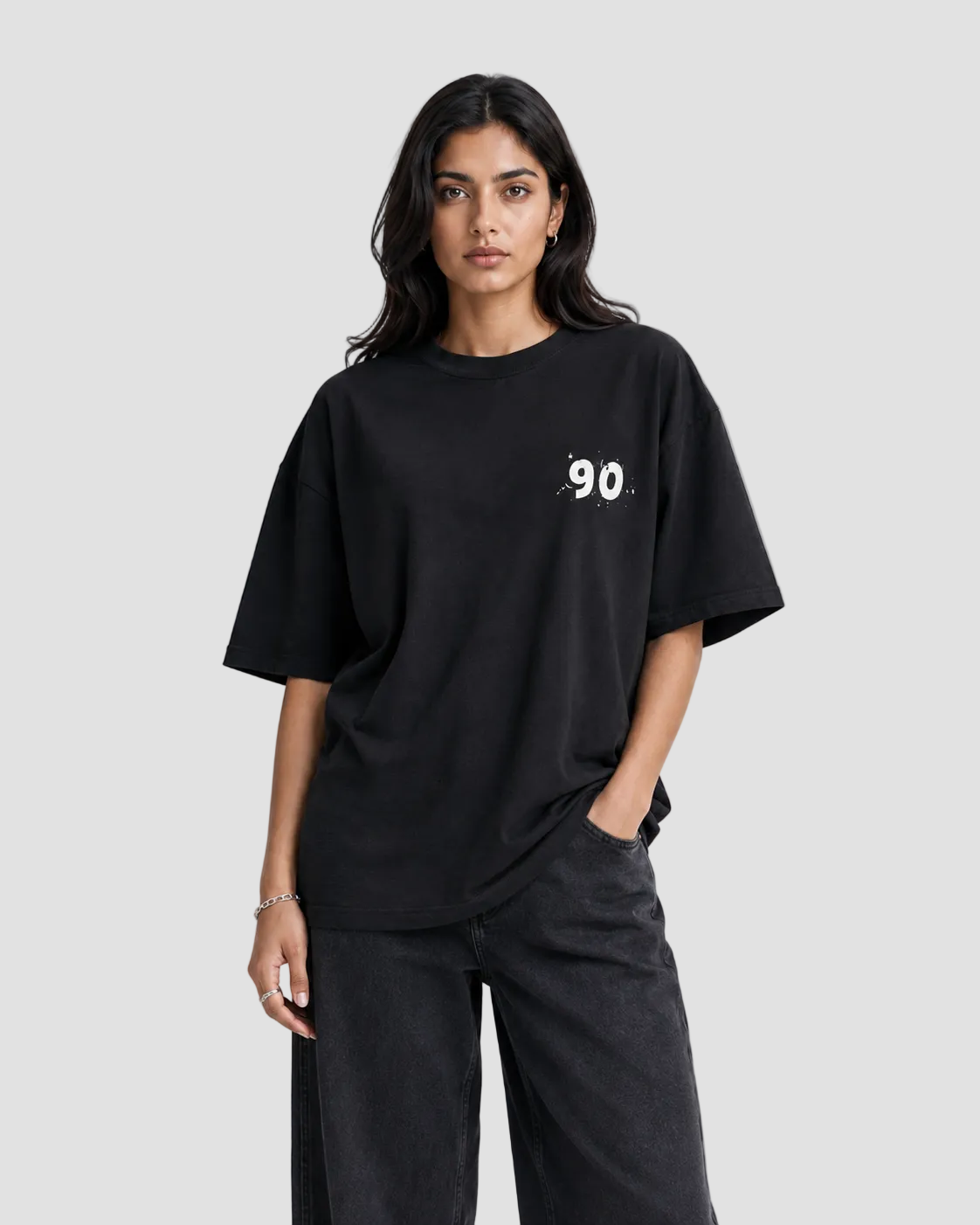 A woman modeling an oversized black t-shirt with a small white graphic print on the left chest, paired with dark charcoal wide-leg pants, against a plain light gray studio background.