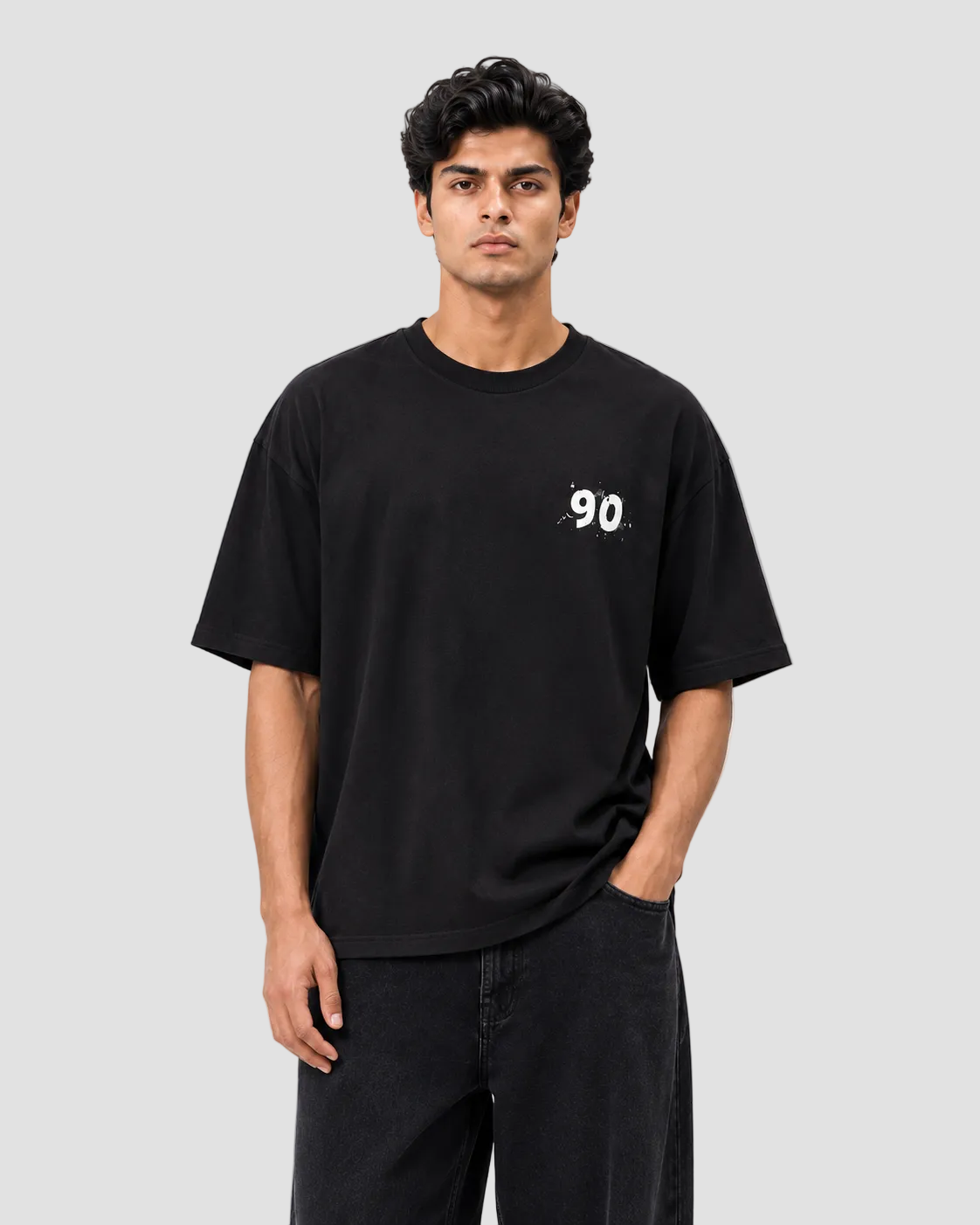 A man wearing a black short-sleeve t-shirt with a small white graphic on the left chest, paired with black jeans, against a plain light gray studio background.