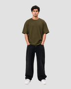 A man wearing an oversized olive green t-shirt, loose black corduroy pants, and white sneakers, standing against a plain light gray studio background.